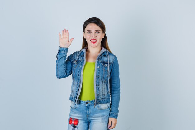 Hermosa joven saludando con la mano abierta en traje de mezclilla y mirando alegre, vista frontal.