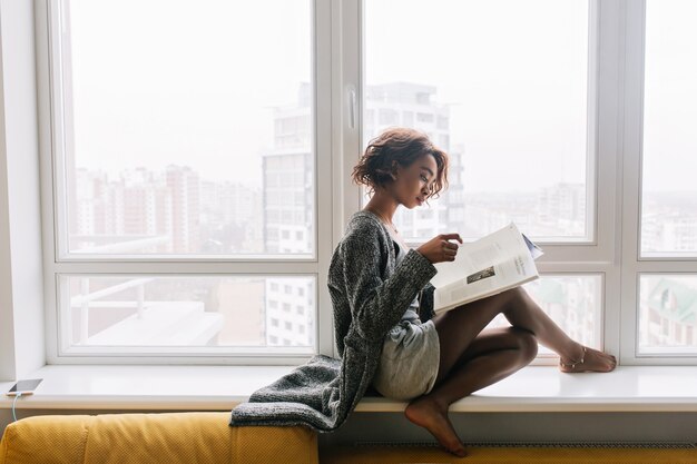 Hermosa joven sentada en el alféizar de la ventana, gran ventana blanca, leyendo una revista, libro. Adolescente africana tiene el pelo corto y rizado, brazalete de oro en la pierna, chaqueta de punto gris larga y pantalones cortos.