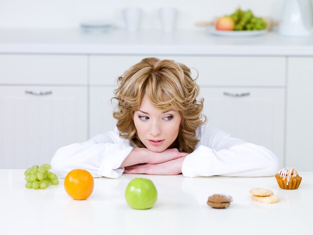 Hermosa joven sentada en la cocina y elegir entre frutas saludables y deliciosos pasteles