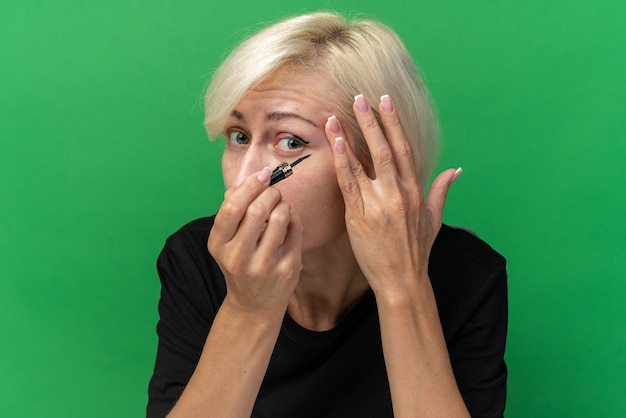 Foto gratuita hermosa joven se sienta a la mesa con herramientas de maquillaje aplicando rímel aislado en la pared verde