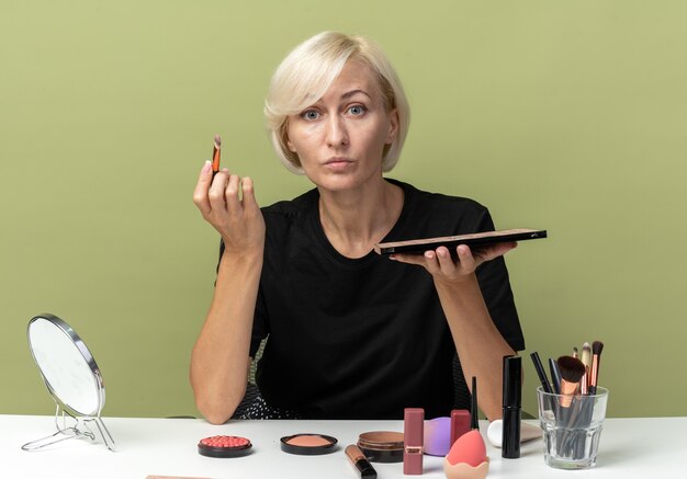 Hermosa joven se sienta a la mesa con herramientas de maquillaje aplicando sombra de ojos con pincel de maquillaje aislado en la pared verde oliva