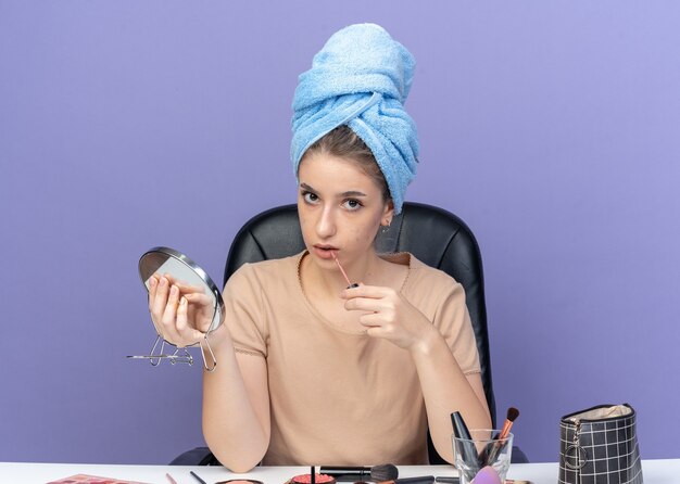 Hermosa joven se sienta a la mesa con herramientas de maquillaje envuelto el cabello en una toalla sosteniendo el espejo aplicando brillo de labios aislado en la pared azul