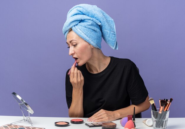 Hermosa joven se sienta a la mesa con herramientas de maquillaje limpiando el cabello con una toalla aplicando lápiz labial aislado en la pared azul