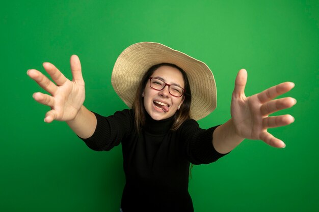 Hermosa joven con sombrero de verano en un cuello alto negro y gafas sonriendo alegremente haciendo gesto de bienvenida