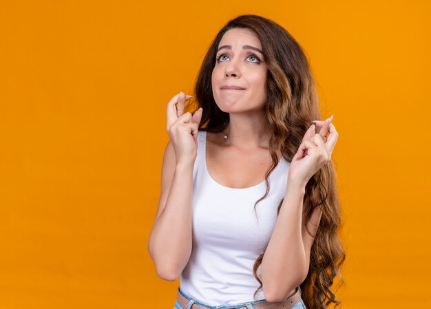 Hermosa joven soñadora mirando hacia arriba y haciendo gestos con los dedos cruzados en el espacio naranja aislado con espacio de copia