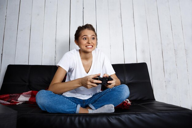 Hermosa joven sonriendo, jugando videojuegos, sentado en el sofá en casa. Copie el espacio.
