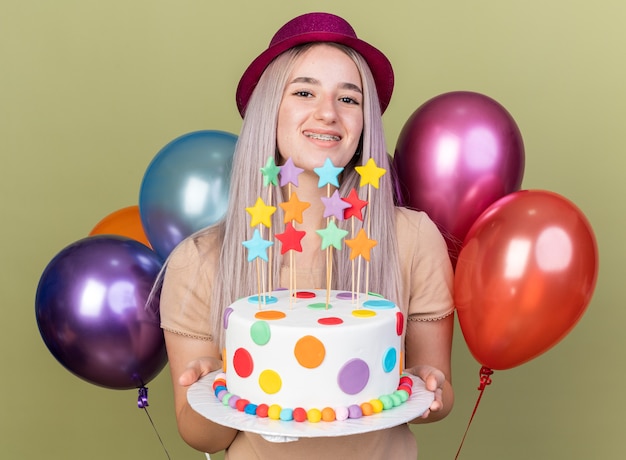 Hermosa joven sonriente con aparatos dentales con gorro de fiesta sosteniendo pastel de pie delante de globos