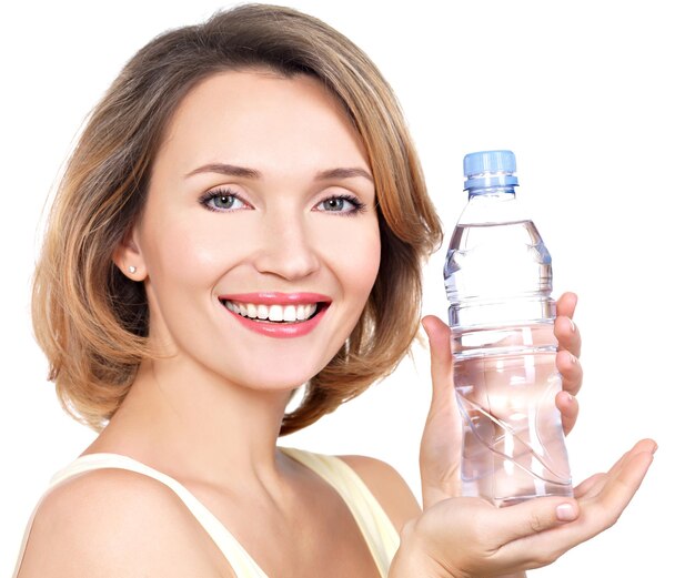 Hermosa joven sonriente con una botella de agua en una pared blanca.