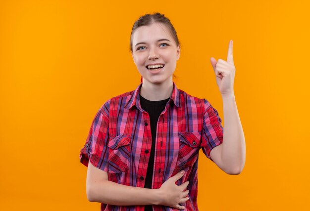 Hermosa joven sonriente con camisa roja apunta con el dedo hacia arriba sobre fondo amarillo aislado