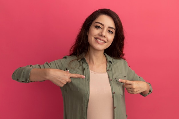 Foto gratuita hermosa joven sonriente con camiseta verde oliva apunta a sí misma aislada en la pared rosa