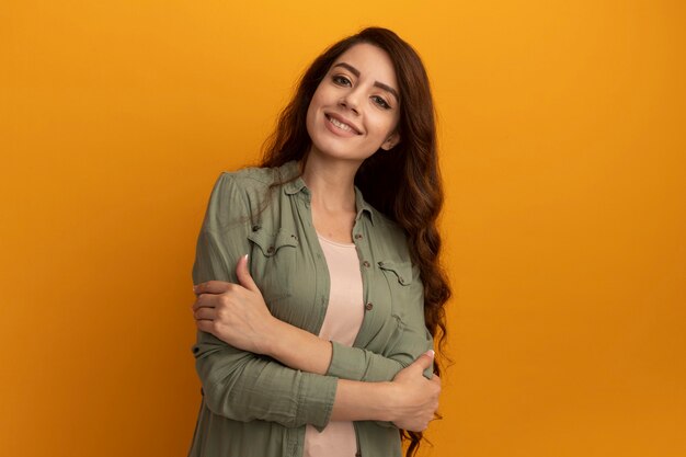 Hermosa joven sonriente con camiseta verde oliva cruzando las manos aisladas en la pared amarilla