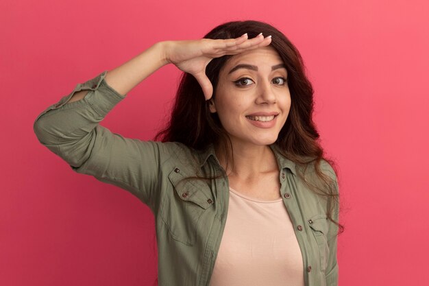 Hermosa joven sonriente con camiseta verde oliva mostrando gesto de saludo aislado en la pared rosa