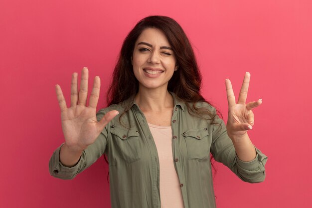 Hermosa joven sonriente con camiseta verde oliva que muestra diferentes números aislados en la pared rosa