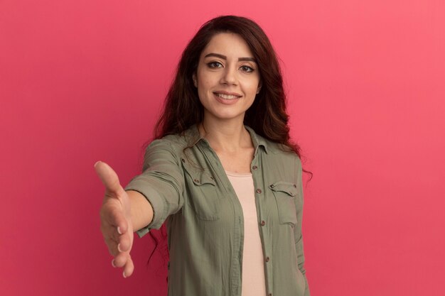 Hermosa joven sonriente con camiseta verde oliva sosteniendo la mano aislada en la pared rosa