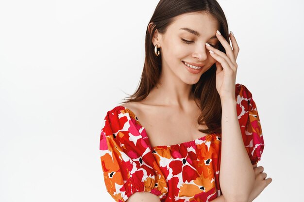 Hermosa joven sonriente coqueta tocándose la cara y mirando hacia abajo tímida sonrojándose de pie en un vestido floral contra el fondo blanco.