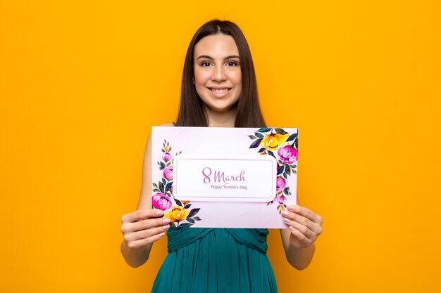 Hermosa joven sonriente en el día de la mujer feliz sosteniendo una postal aislada en la pared naranja