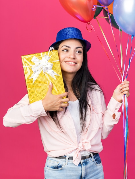 Hermosa joven sonriente con gorro de fiesta sosteniendo globos con caja de regalo