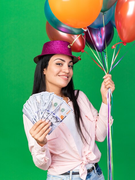 Hermosa joven sonriente con gorro de fiesta sosteniendo globos con dinero en efectivo