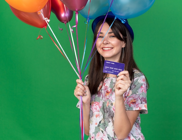 Hermosa joven sonriente con gorro de fiesta sosteniendo globos con tarjeta de crédito