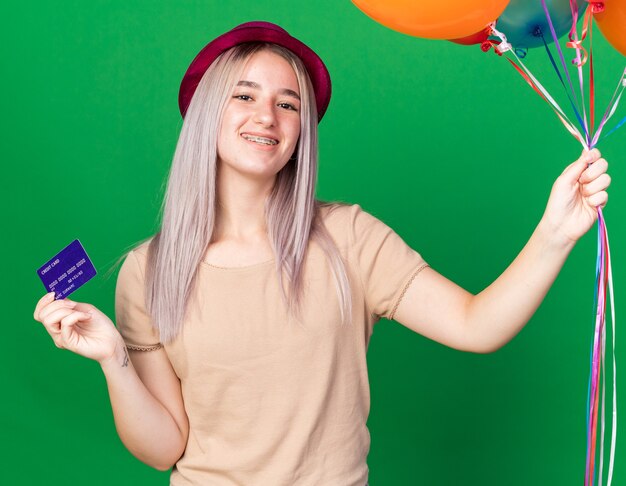 Hermosa joven sonriente con gorro de fiesta y tirantes sosteniendo globos con tarjeta de crédito