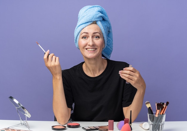 Hermosa joven sonriente se sienta a la mesa con herramientas de maquillaje limpiando el cabello con una toalla sosteniendo brillo de labios aislado sobre fondo azul.