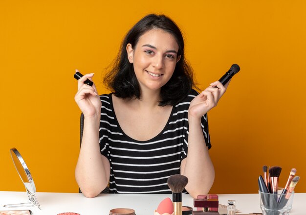 Hermosa joven sonriente se sienta a la mesa con herramientas de maquillaje con pincel en polvo aislado sobre fondo naranja