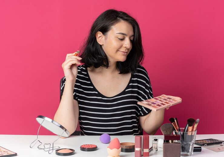 Foto gratuito hermosa joven sonriente se sienta a la mesa con herramientas de maquillaje sosteniendo y mirando la paleta de sombra de ojos con pincel de maquillaje aislado sobre fondo rosa