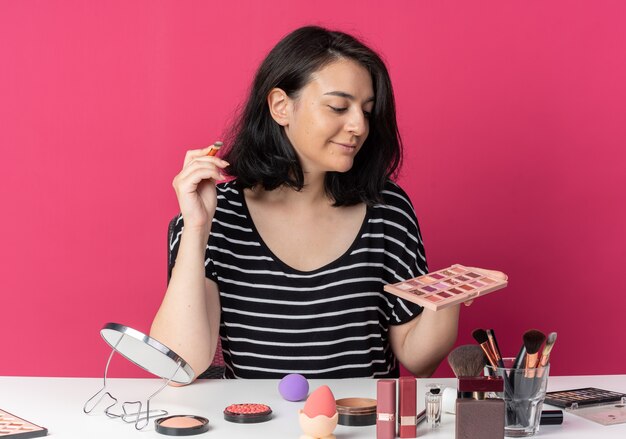 Hermosa joven sonriente se sienta a la mesa con herramientas de maquillaje sosteniendo y mirando la paleta de sombra de ojos con pincel de maquillaje aislado sobre fondo rosa