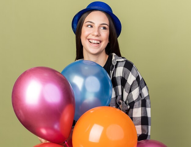 Hermosa joven sonriente con sombrero azul de pie detrás de globos