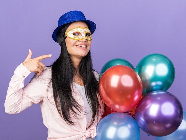 Hermosa joven sonriente con sombrero de fiesta y antifaz de mascarada sosteniendo globos apunta a sí misma aislada en la pared azul