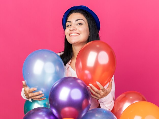 Hermosa joven sonriente con sombrero de fiesta de pie detrás de globos