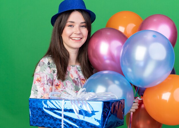 Hermosa joven sonriente con sombrero de fiesta sosteniendo globos con caja de regalo aislada en la pared verde