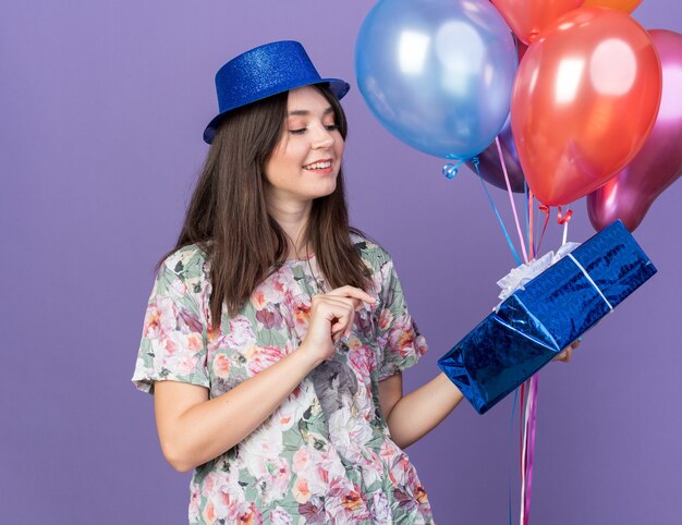 Hermosa joven sonriente con sombrero de fiesta sosteniendo y mirando globos con caja de regalo