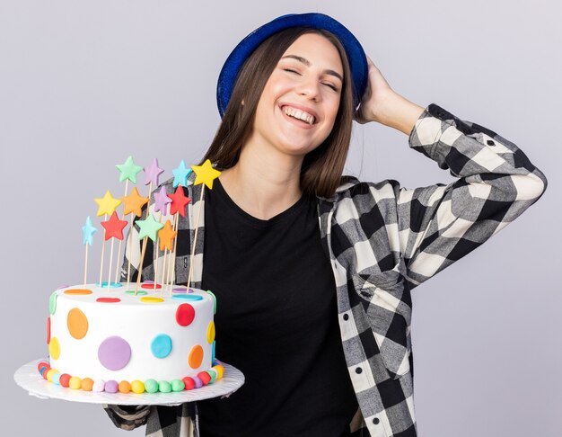 Hermosa joven sonriente con sombrero de fiesta sosteniendo la torta poniendo la mano en la cabeza aislada en la pared blanca