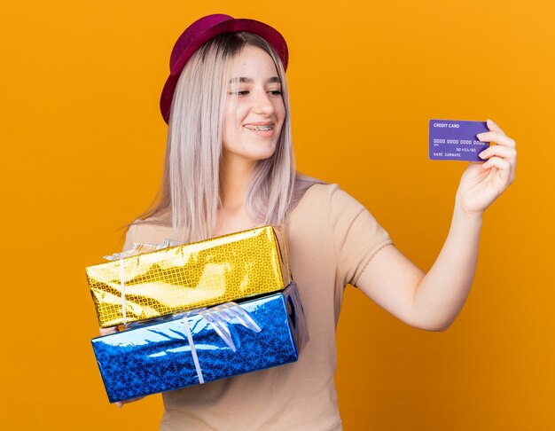 Hermosa joven sonriente con sombrero de fiesta con tirantes sosteniendo cajas de regalo y mirando la tarjeta de crédito en su mano aislada en la pared naranja