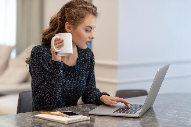 Hermosa joven sonriente trabajando en la computadora portátil y tomando café mientras está sentado en casa