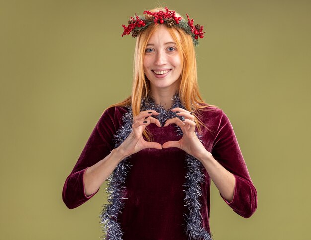 Hermosa joven sonriente con vestido rojo con corona y guirnalda en el cuello mostrando gesto de corazón aislado sobre fondo verde oliva