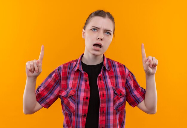 Hermosa joven sorprendida con camisa roja señala con el dedo hacia arriba sobre fondo amarillo aislado