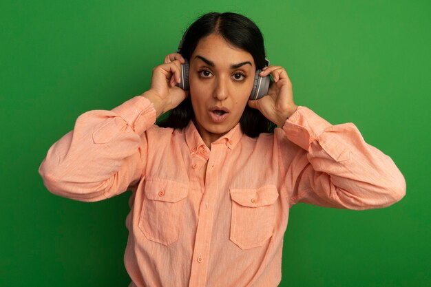 Foto gratuita hermosa joven sorprendida con camiseta rosa con auriculares aislados en la pared verde