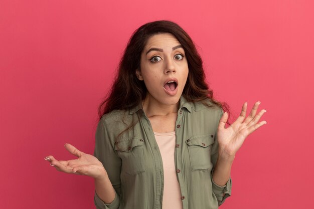 Hermosa joven sorprendida con camiseta verde oliva extendiendo las manos aisladas en la pared rosa