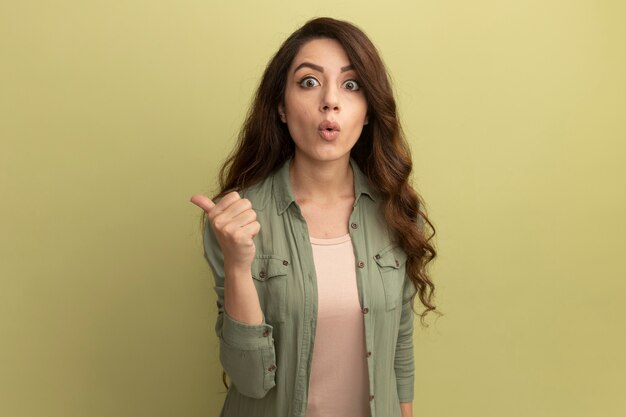 Hermosa joven sorprendida con camiseta verde oliva que muestra el pulgar hacia arriba aislado en la pared verde oliva