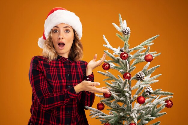 Hermosa joven sorprendida de pie cerca del árbol de Navidad con sombrero de Navidad aislado sobre fondo naranja