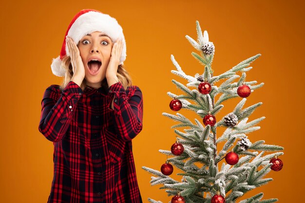 Hermosa joven sorprendida de pie cerca del árbol de Navidad con sombrero de Navidad poniendo las manos en las mejillas aisladas sobre fondo naranja