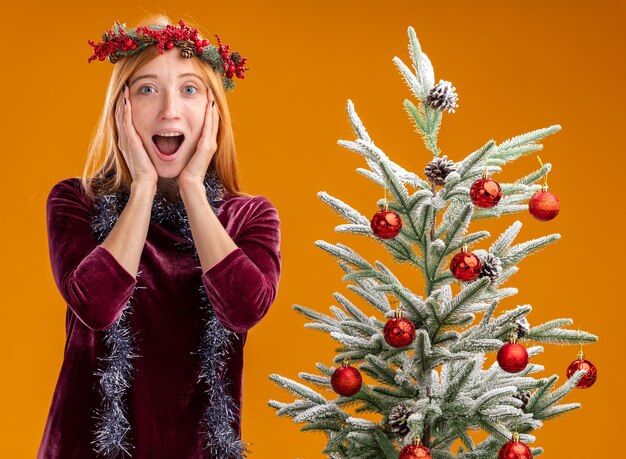 Hermosa joven sorprendida de pie cerca del árbol de Navidad con vestido rojo y corona con guirnalda en el cuello poniendo las manos en las mejillas aisladas sobre fondo naranja