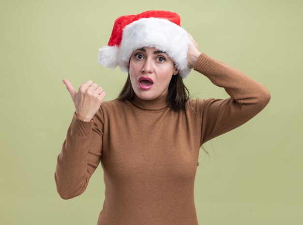 Hermosa joven sorprendida con puntos de sombrero de navidad en el lado aislado sobre fondo verde oliva con espacio de copia