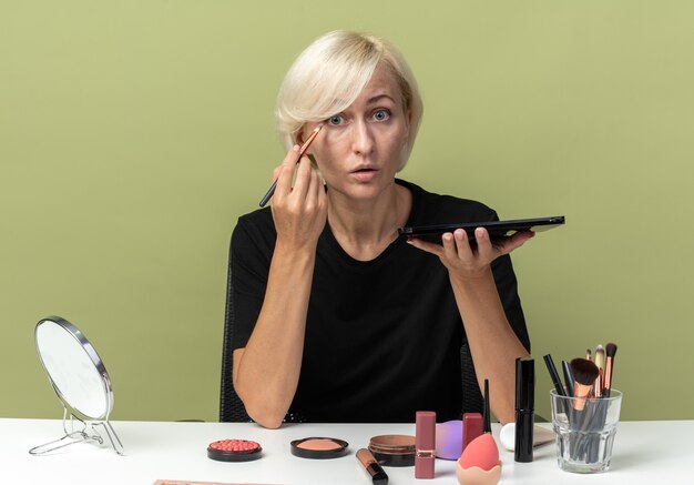 Hermosa joven sorprendida se sienta a la mesa con herramientas de maquillaje aplicando sombra de ojos con pincel de maquillaje aislado sobre fondo verde oliva