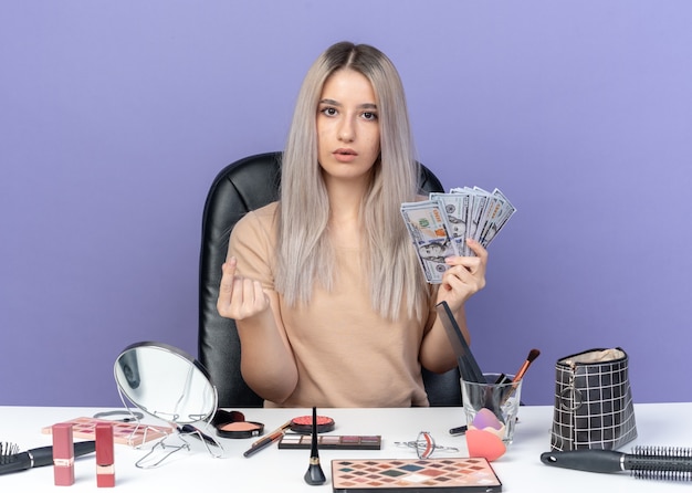 Hermosa joven sorprendida se sienta a la mesa con herramientas de maquillaje con dinero en efectivo mostrando gesto de propina aislado sobre fondo azul.