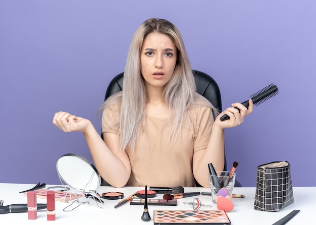 Hermosa joven sorprendida se sienta a la mesa con herramientas de maquillaje con peine aislado sobre fondo azul.