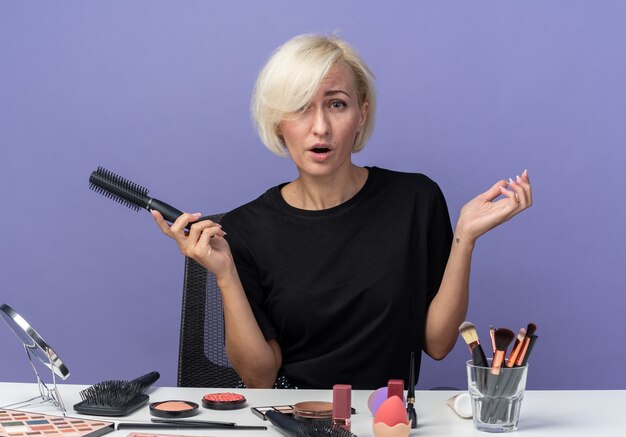 Hermosa joven sorprendida se sienta a la mesa con herramientas de maquillaje con peine aislado sobre fondo azul.