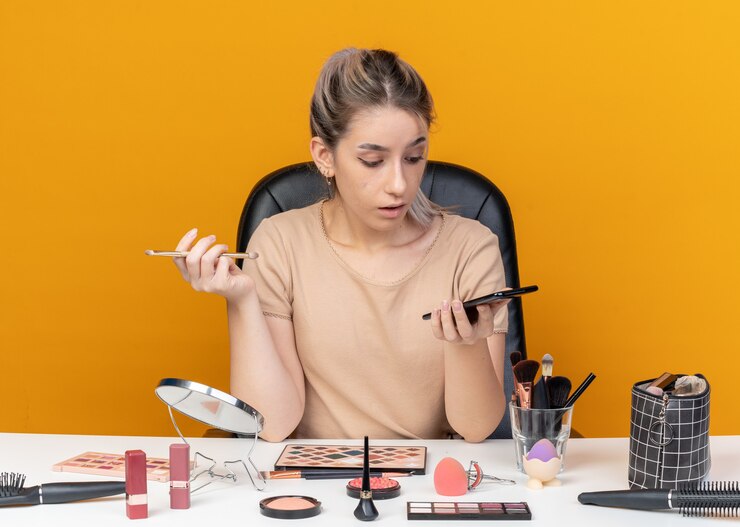 Foto gratuito hermosa joven sorprendida se sienta a la mesa con herramientas de maquillaje sosteniendo pincel de maquillaje y mirando el teléfono en su mano aislado sobre fondo naranja